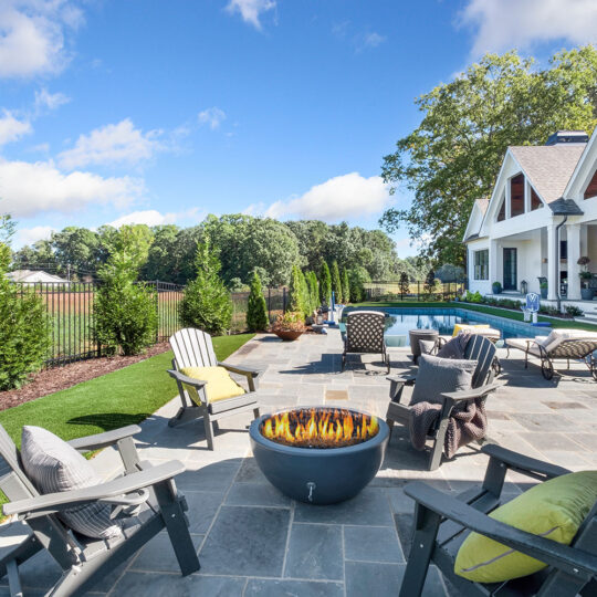 Spacious backyard with a stone patio featuring a central fire pit surrounded by chairs, a swimming pool, and a modern white house with large windows and a manicured lawn under a blue sky.