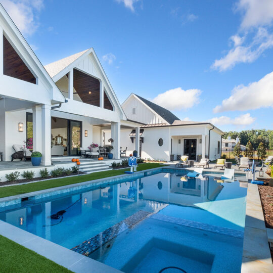 Modern white house with a spacious patio overlooking a rectangular pool. The pool area is surrounded by landscaping and outdoor furniture. The sky is clear with a few clouds, creating a serene and inviting atmosphere.