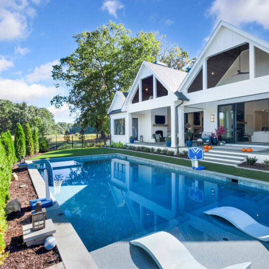 Modern white house with large windows and gabled roof, featuring an outdoor pool with two white loungers partially submerged. The poolside area has potted plants and a basketball hoop. Trees and a grassy field are visible in the background.