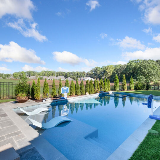 A luxurious outdoor swimming pool with lounge chairs on a stone deck. The pool area is surrounded by lush greenery and a metal fence. Beyond the fence, there is a field with trees under a blue sky with scattered clouds.