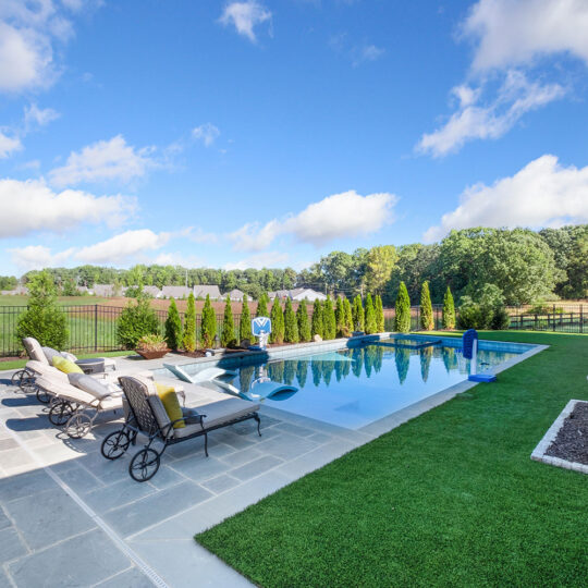 Luxurious backyard with a rectangular pool, surrounded by paved stone tiles and manicured grass. Lounge chairs line one side, while a row of small trees borders the pool. A clear blue sky with fluffy clouds completes the serene setting.