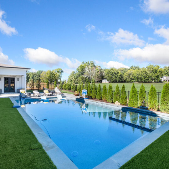 A modern backyard features a clear blue swimming pool surrounded by artificial grass. Tall shrubs line the fence, with a white house and patio area with outdoor furniture nearby. A vast, green field stretches beyond the pool area under a blue sky.