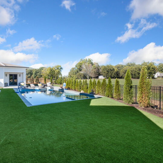 Modern house exterior with a pool surrounded by lush green lawn and trees. The sky is blue with scattered clouds. A person stands near the pool, and theres a fenced area with more trees beyond.