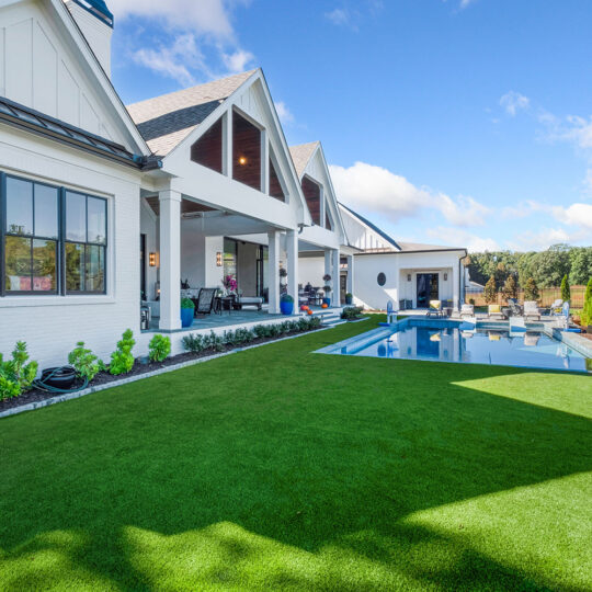 Spacious backyard featuring a modern white house with large windows, neatly trimmed green lawn, and an inviting swimming pool. There are several lounge chairs around the pool, and the sky is clear and blue.