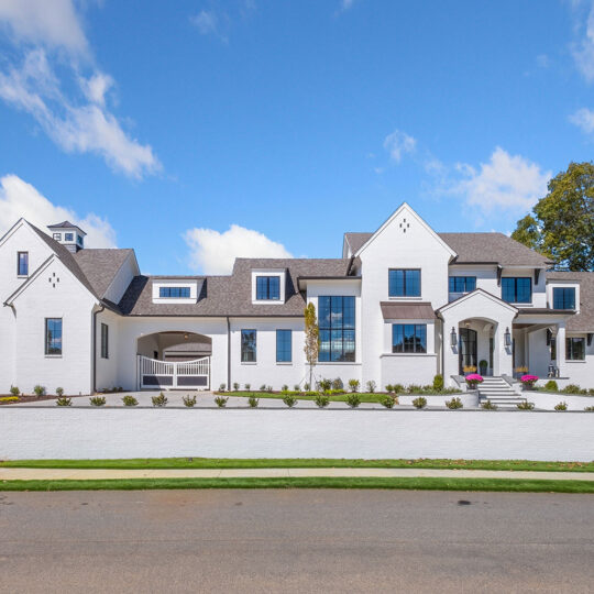 A large, modern white house with multiple gables and large windows under a clear blue sky. A manicured lawn and small shrubs line the front yard, and there’s a paved driveway on the left side.