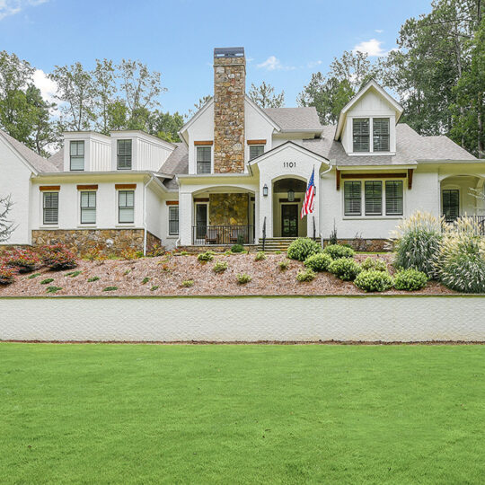 A large, two-story house with white exteriors, gabled roofs, and a prominent chimney. A well-maintained yard with a trimmed lawn and landscaped bushes surrounds the home. An American flag hangs on the front porch. Trees provide a backdrop.