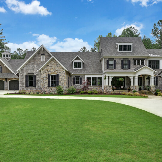 Large stone house with gray roof, multiple dormer windows, and a three-car garage. The home is surrounded by lush green grass and tall trees. The driveway curves in front of the entrance, and the sky is clear with fluffy clouds.