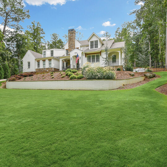 A large house with a steeply pitched roof sits on a landscaped lawn surrounded by trees. A winding gravel driveway leads to the front entrance, and the property is elevated with stone retaining walls adorned with shrubs.