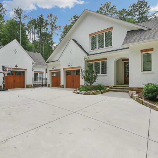 A large white house with a modern design featuring multiple gables and large wooden garage doors. Theres a basketball hoop on the left side of the spacious driveway. Trees are in the background under a partly cloudy sky.