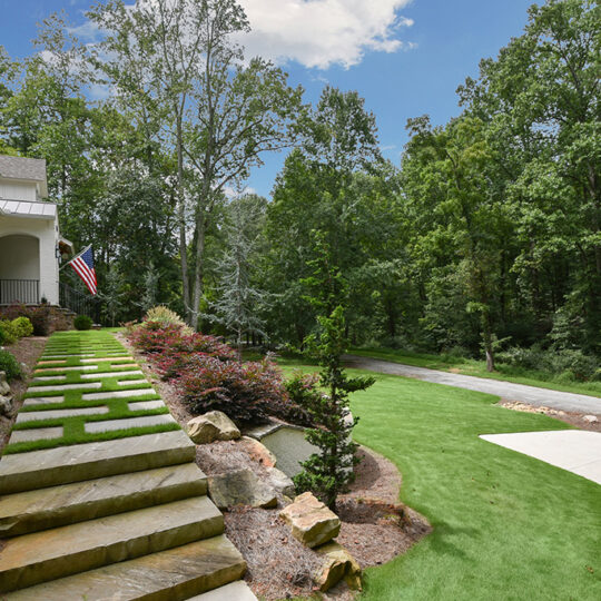 Stone steps with grass insets lead to a house surrounded by lush greenery. An American flag is displayed on the porch. A curved driveway and a forested area are visible under a clear blue sky.