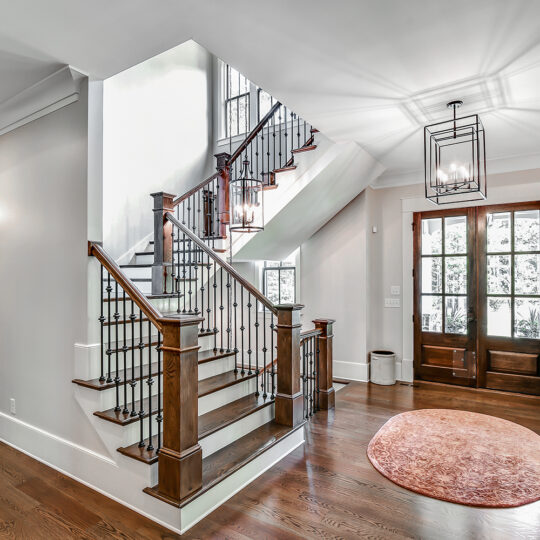 A spacious foyer with wooden flooring and a staircase featuring wooden handrails and black spindles. Theres a round rug on the floor, a large window by the entrance, and a decorative mirror on the left wall.