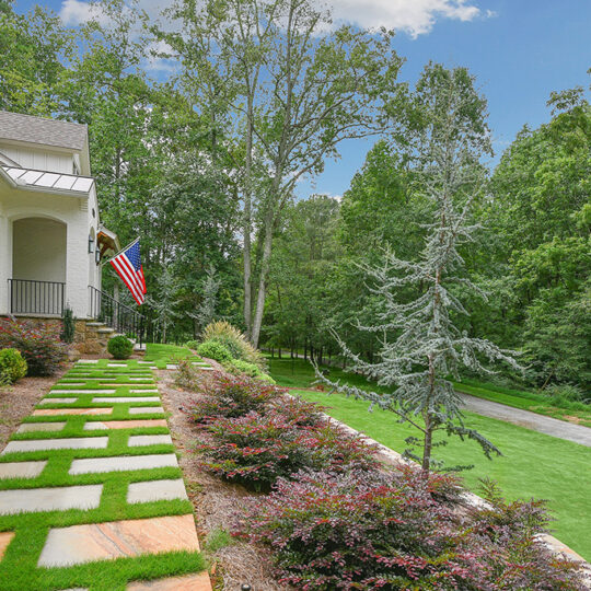 Front yard of a house with a modern stone walkway surrounded by green grass and shrubs. An American flag hangs from the porch. Mature trees and a lush forest are in the background under a partly cloudy sky.