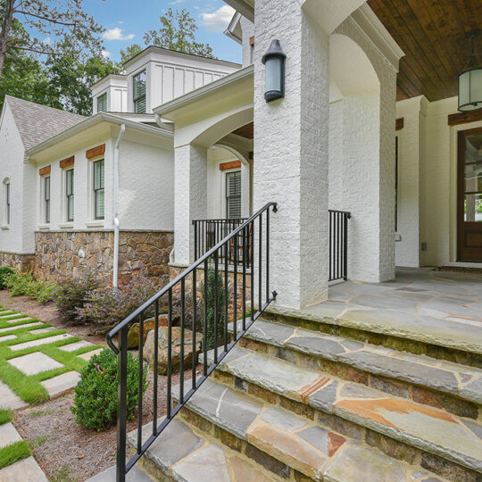 A modern house entrance with white brick walls and stone steps leading up to a wooden front door. Theres a black metal railing and a path with stone tiles set in grass. Two wall lanterns are mounted near the entrance. Trees are visible in the background.