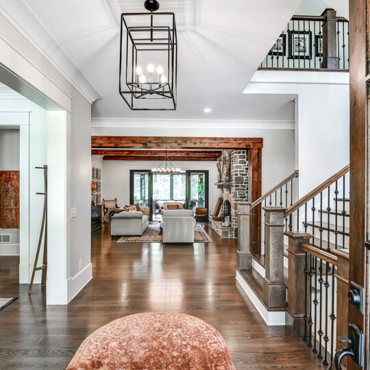 Spacious living room with hardwood floors, visible staircase, and elegant chandelier. The room features a seating area with sofas, a stone fireplace, and large windows offering natural light. Decor includes rugs and a mix of wooden and modern elements.