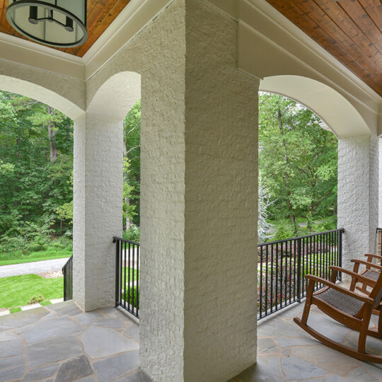 Covered porch with a stone floor, white arches, and a wooden ceiling. Two wooden rocking chairs face a lush green yard with a road running along the side.