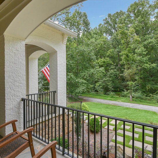 A cozy porch features two wooden chairs with a view of a lush, green yard and a flagpole displaying the American flag. The porch has a white brick wall and a black metal railing, opening to a paved pathway and dense trees in the background under a clear sky.