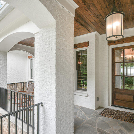 Covered porch with white brick walls and stone flooring. Features a wooden door with glass panels, a black railing, and a large pendant light hanging from a wood-paneled ceiling. Several archways and a welcome mat are visible.