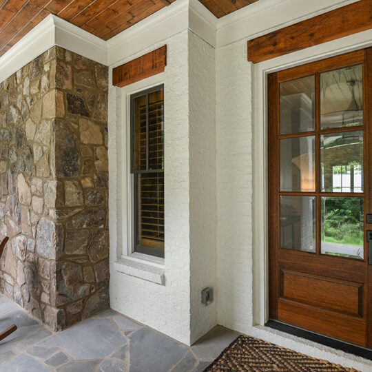 A cozy porch features a wooden door with glass panels, a stone wall, and two wooden rocking chairs on a stone floor. A doormat is placed near the door, and a window with shutters is next to it. A view of greenery is visible through the door.