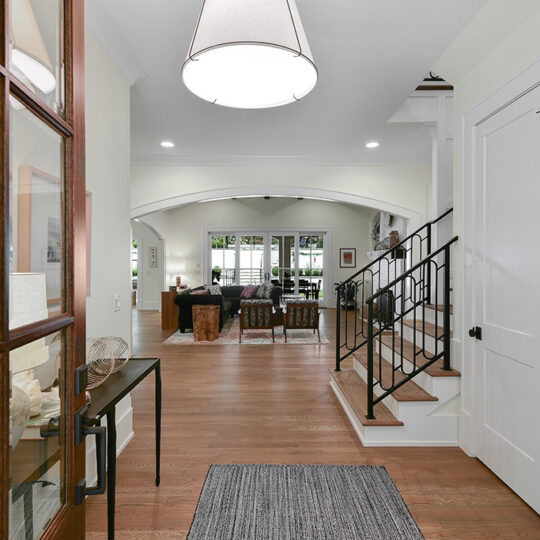 Spacious homes entryway featuring hardwood floors. A door with glass panels opens to a room with a rug, stairs on the right, and a sitting area with a couch visible through an archway. Large windows let in natural light, and a modern light fixture hangs above.