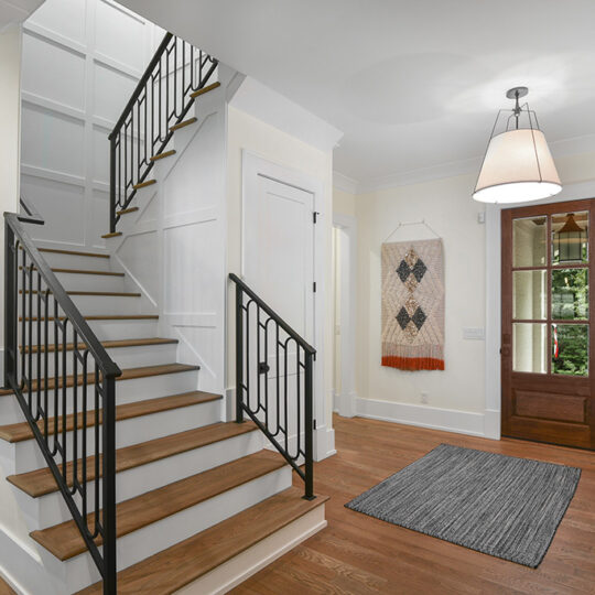 A hallway with wooden floors featuring a wooden staircase with black metal railings. To the right, there is a small table with a lamp, a rug, a wall hanging with geometric patterns, and a door with glass panels.