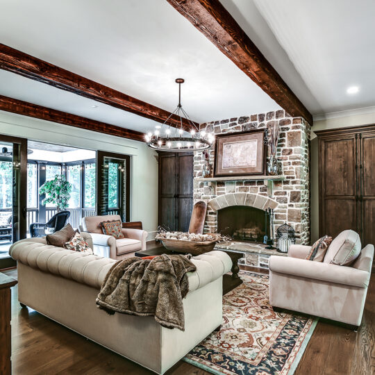 A cozy living room with a stone fireplace, wooden beams on the ceiling, and hardwood floors. Theres a light-colored sectional sofa, armchairs, a patterned rug, and a large chandelier. Glass doors lead to an outdoor space with trees visible.