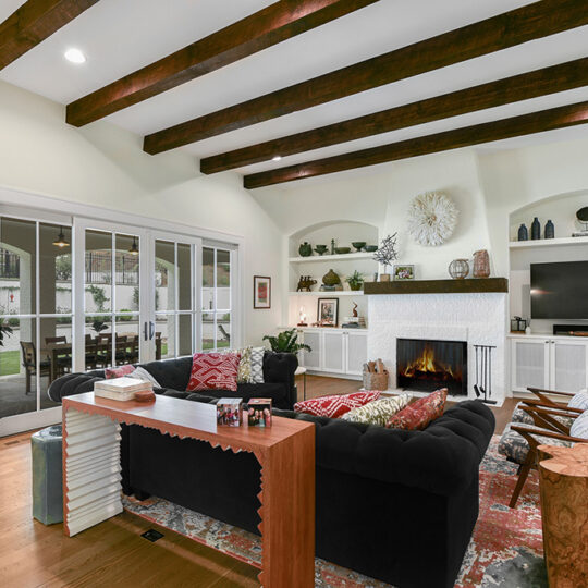 A cozy living room with exposed wooden beams and a large brick fireplace. Theres a black sectional sofa with colorful cushions, a wooden coffee table, and built-in shelves around the fireplace. Large glass doors lead to an outdoor area.