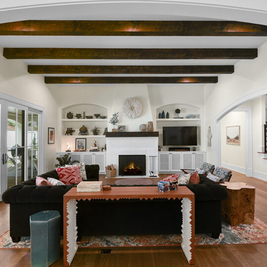 Spacious living room with a black sofa, wooden beams on the ceiling, and a fireplace. A flat-screen TV sits above the fireplace. Sliding glass doors lead to an outdoor area. A staircase is visible to the right. Decor includes art pieces and vases.