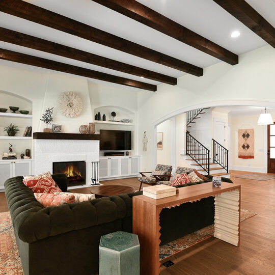 A cozy living room features a dark green sectional sofa, a fireplace, and a flat-screen TV. Wooden beams line the ceiling, and built-in shelves display decorative items. A staircase and hallway are visible in the background.