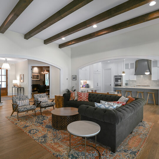 Spacious living room with a large gray sectional sofa, patterned rug, and two armchairs. Exposed wooden beams adorn the ceiling. An open kitchen is visible in the background. There’s a staircase with a black railing to the left.