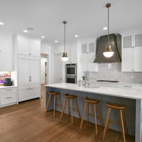 Modern kitchen with white cabinets, gray island, and wooden stools. Pendant lights hang above the island. A built-in desk with a chair and a bulletin board is visible to the left. Stainless steel appliances and a range hood are present.