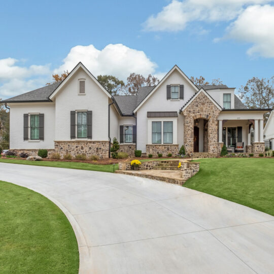 A large, modern house with a stone and brick facade stands on a green lawn. It features multiple peaked roofs and shuttered windows. A curved concrete driveway leads to the entrance. The sky is partly cloudy.