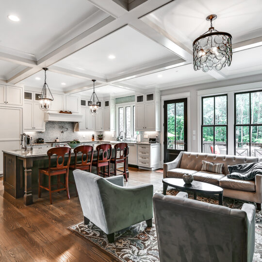 Spacious open-concept living area featuring a modern kitchen with a central island and wooden bar stools. Adjacent is a cozy seating area with sofas and a table. Large windows offer a view of greenery outside, and elegant lighting fixtures hang from the ceiling.