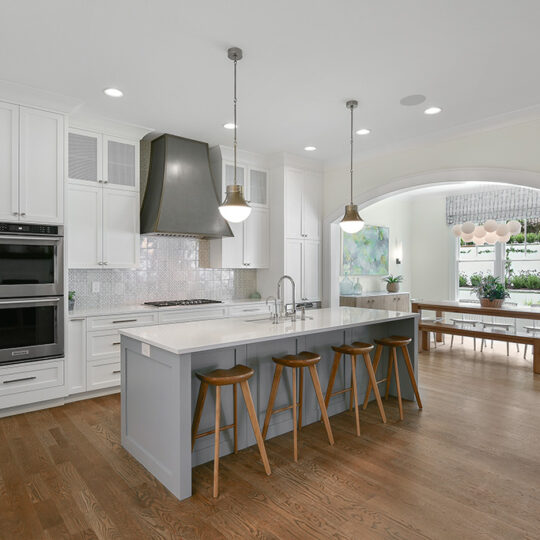 Modern kitchen with a large gray island and four wooden stools. It features stainless steel appliances, white cabinets, pendant lights, and a backsplash. An adjacent dining area with a long table is visible through an arched opening.