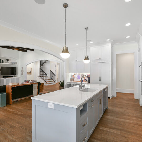 Bright, modern kitchen with a large island featuring a white countertop and gray cabinetry. Pendant lights hang above the island. In the background, theres a living room with a sofa, TV, and a staircase leading upstairs.