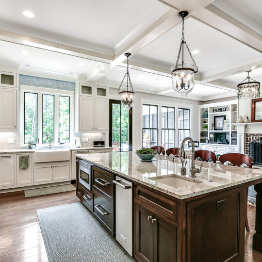 Spacious kitchen with a large marble island and sink, surrounded by white cabinets and stainless steel appliances. Elegant pendant lights hang above the island. A dining area and fireplace can be seen in the adjacent room, with large windows providing natural light.