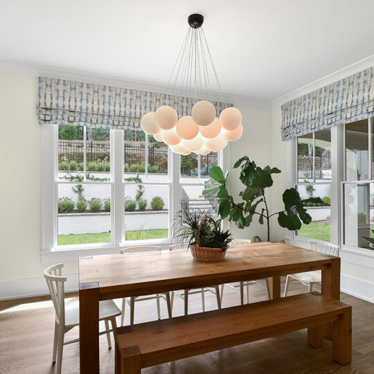 A dining room with a wooden table and mixed chairs under a modern cluster light fixture. Large windows with blue patterned curtains show a garden outside. Plants decorate the table and a sideboard on the left, enhancing the rooms natural feel.