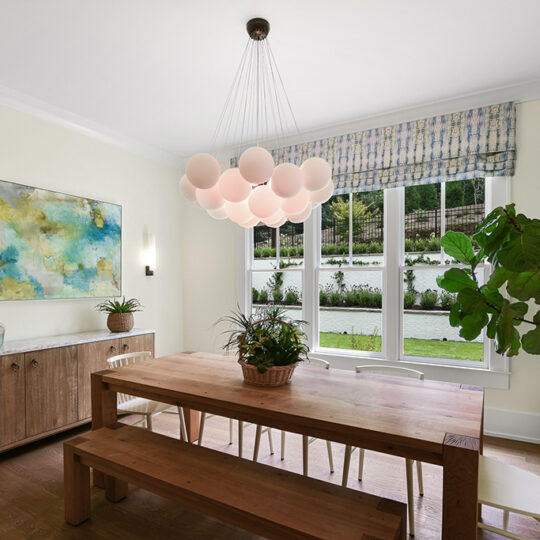 A dining room with a wooden table, benches, and white chairs. A cluster of round pendant lights hangs above. Theres a wooden sideboard with decor items, a large abstract painting, and a tall potted plant near windows showing a garden view.