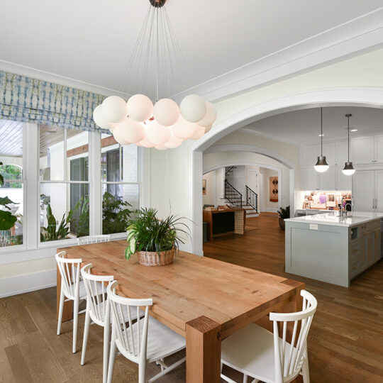 A dining area with a wooden table and white chairs is adorned with a modern, round cluster light fixture. Large windows with patterned curtains illuminate the space. A potted plant decorates the corner, and an arched opening leads to a kitchen.