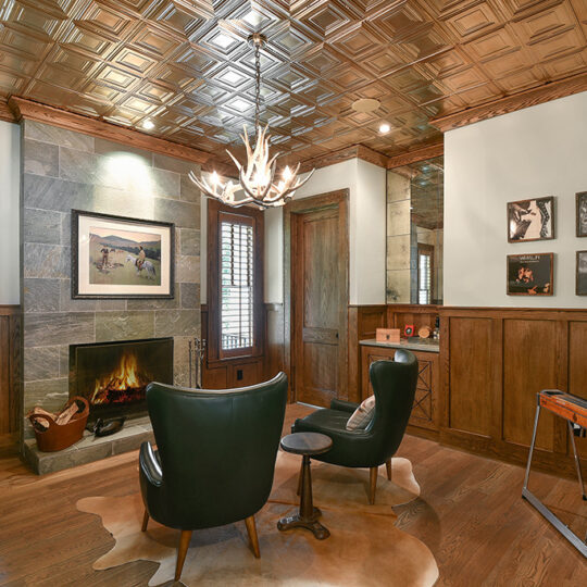 Cozy room with wood paneling, featuring a fireplace, two dark green chairs, and a small table on a cowhide rug. A chandelier with antler-like design hangs from the metallic ceiling. Art and photos adorn the walls, adding a personal touch.