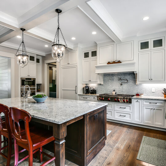 Spacious kitchen featuring a large island with a white marble countertop and dark wooden base. Red chairs are placed around the island. White cabinetry lines the walls, with a stove in the middle. Pendant lights hang from the ceiling, and hardwood floors are visible.