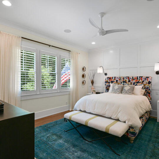 A cozy bedroom with a colorful patterned headboard bed, white bedding, and blue rug. A window with blinds shows a view of greenery. White walls, a ceiling fan, and a side table are also visible. An American flag is partially seen outside.