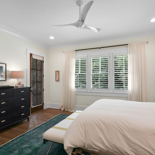 A bedroom with a white bed, green rug, and a dark dresser on the left. A ceiling fan hangs from the ceiling, and a large window with curtains is centered on the back wall. A floor lamp is next to the bed, and the walls are light-colored.