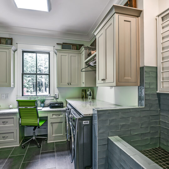 A laundry room with a desk and green chair by a window. The space includes gray cabinets, a washer and dryer, and an industrial-style sink. Shelves hold baskets and decor, and a skylight adds natural light.