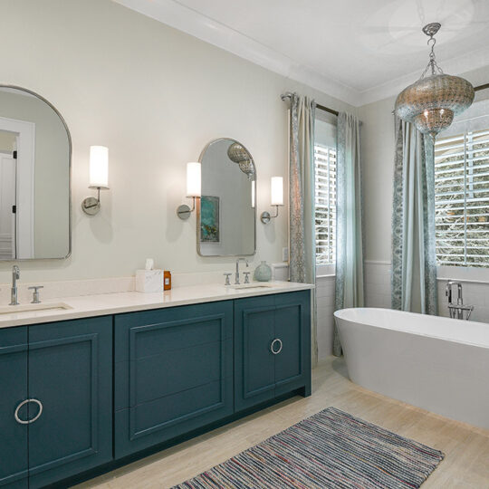 A modern bathroom with a blue double-sink vanity, two oval mirrors, and wall-mounted lights. A freestanding white bathtub sits near a window with gray curtains. A colorful rug is on the floor, and a decorative light fixture hangs from the ceiling.