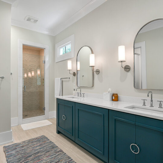 Modern bathroom with a double sink vanity featuring teal cabinets and two oval mirrors. Wall-mounted lights flank the mirrors. A glass-door shower is in the background, and a striped rug lies on the floor. A towel hangs on a nearby rack.