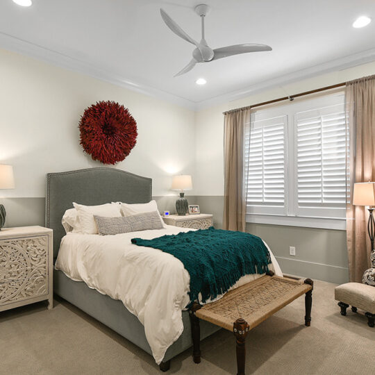 A cozy bedroom with a plush bed, teal throw, and intricate side tables. A red circular wall art decorates the wall above the bed. A patterned armchair and ottoman sit beside a window with beige curtains and blinds. Ceiling fan and lamps are lit.