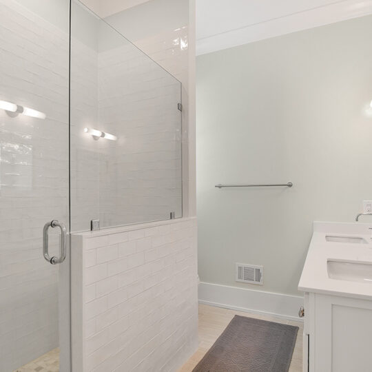 A modern bathroom with a glass-enclosed shower and white tile walls. It features a double vanity with two sinks and mirrors, and soft lighting on light green walls. A gray rug is on the light wood floor.