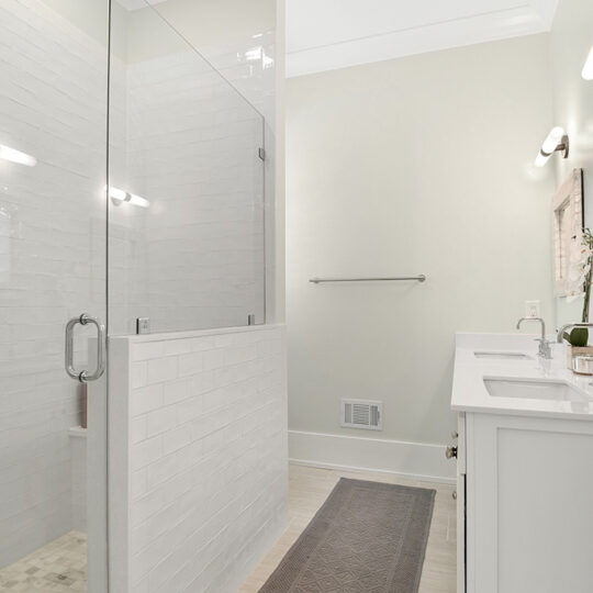 A modern bathroom with a glass-enclosed shower on the left, featuring white subway tiles. To the right, a white vanity with a double sink and two wall-mounted lights above. A gray rug lies on the beige tiled floor.