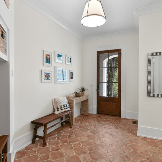 A cozy entryway with terracotta tiles, a wooden bench, and a mirror on the right wall. Photos hang on the left wall above the bench, and a wooden door with a glass window is in the center, flanked by plants on a small table.