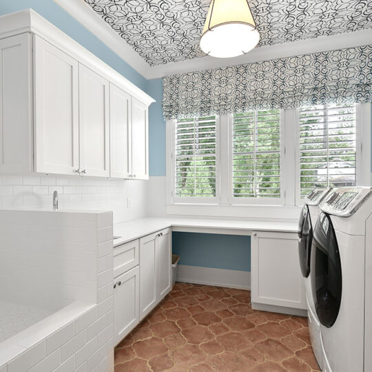 Laundry room with light blue walls, white cabinets, and a patterned black and white ceiling. There are two front-loading washing machines, white countertops, a built-in sink, and large windows with white shutters and a floral valance.